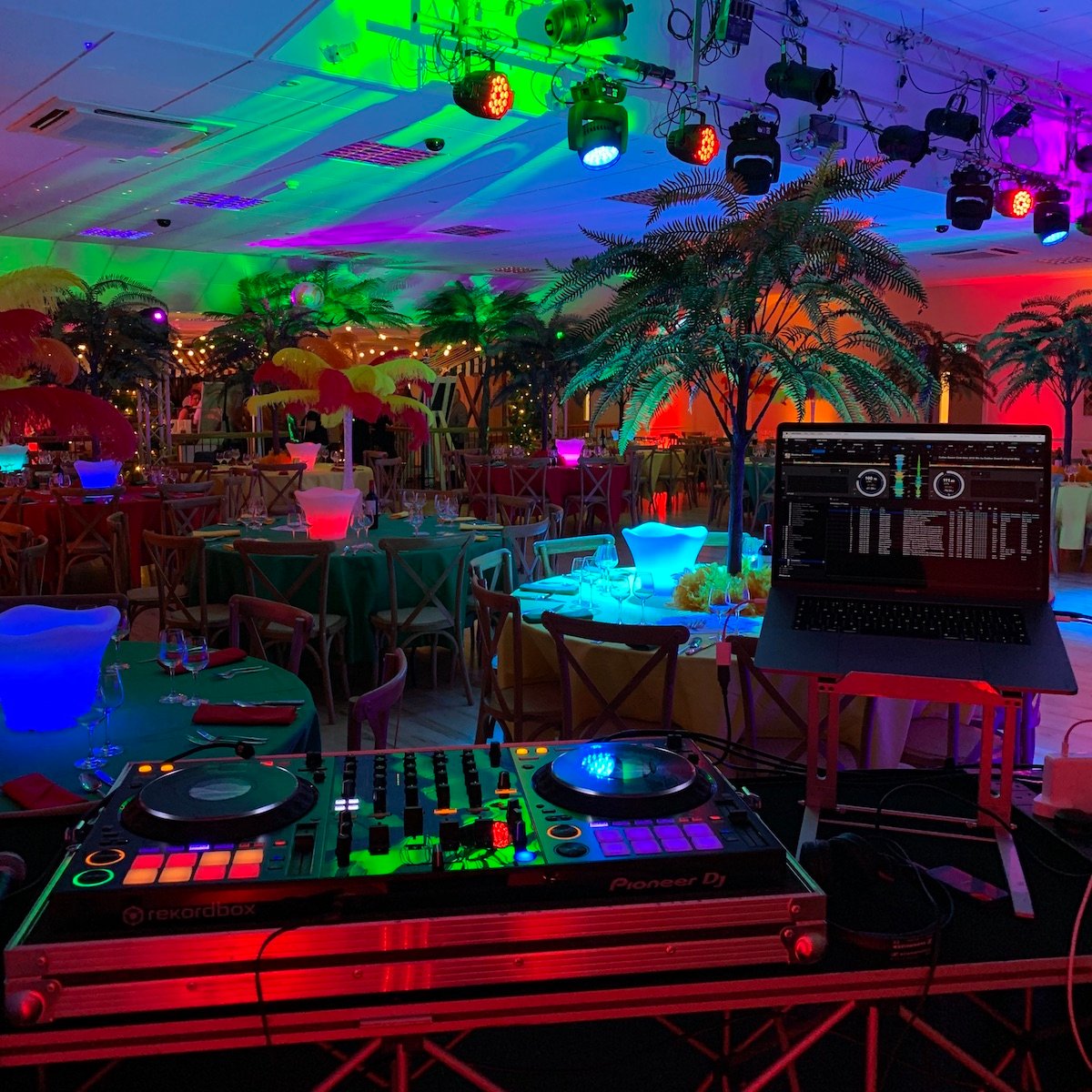 Picture of room decorated for a tropical theme DJ party. Showing palm trees, LED ice buckets, a Pioneer DDJ-1000 DJ controller and a MacBook Pro running RekordBox software.